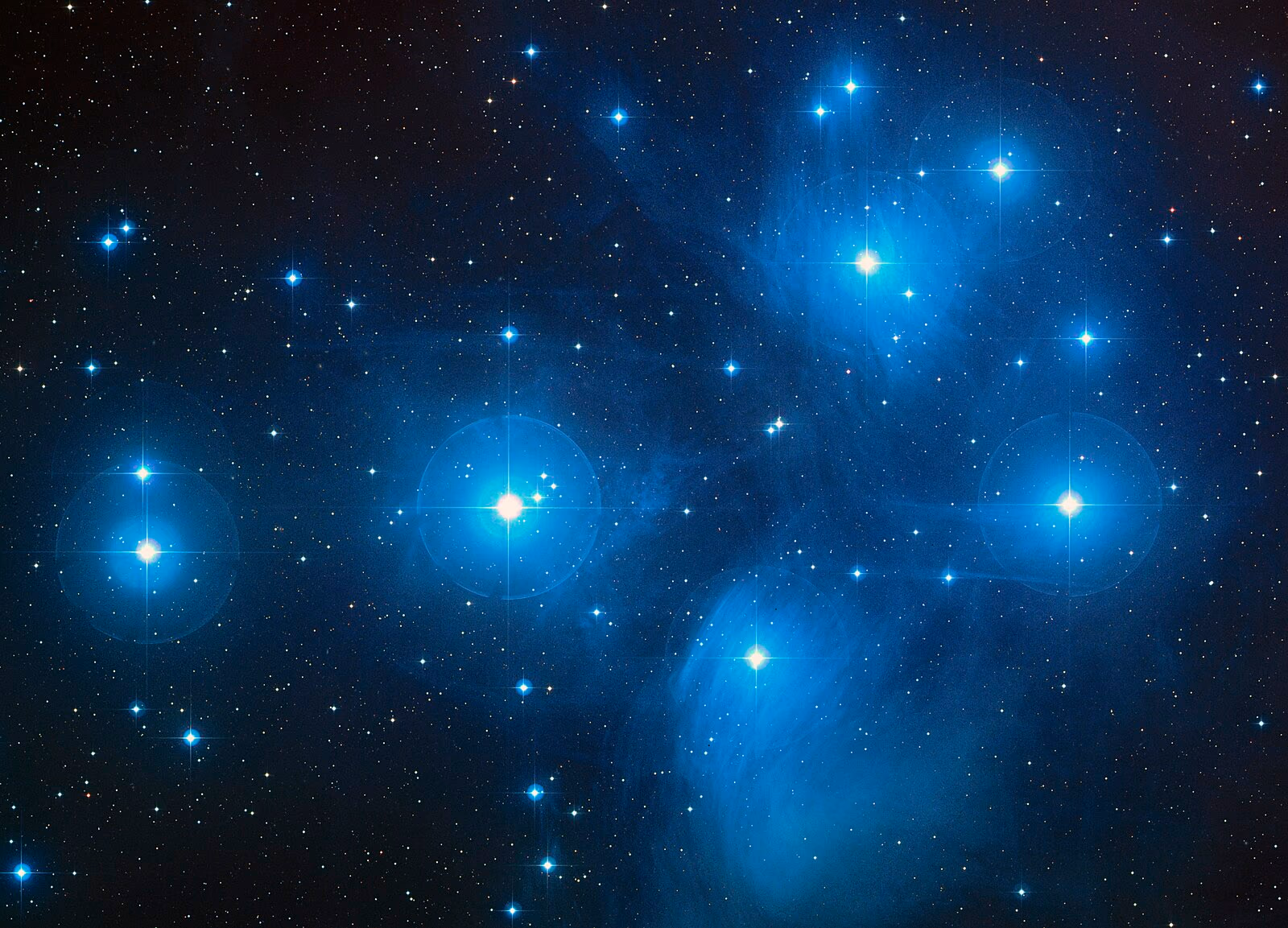The Pleiades star cluster glowing blue with wispy reflection nebulae against a dark star field.