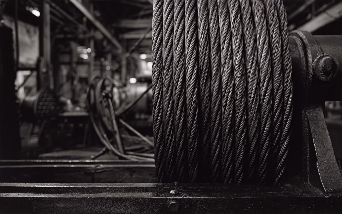 A large format black and white photograph of heavy steel cable wound tightly on a drum, every strand resolved in sharp focus, the machine shop behind it falling away into shallow depth of field.